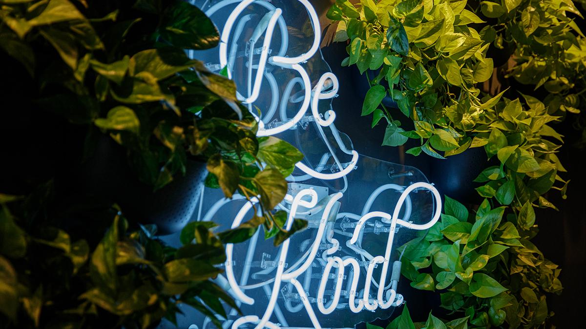Neon sign with 'Be Kind' surrounded by green foliage